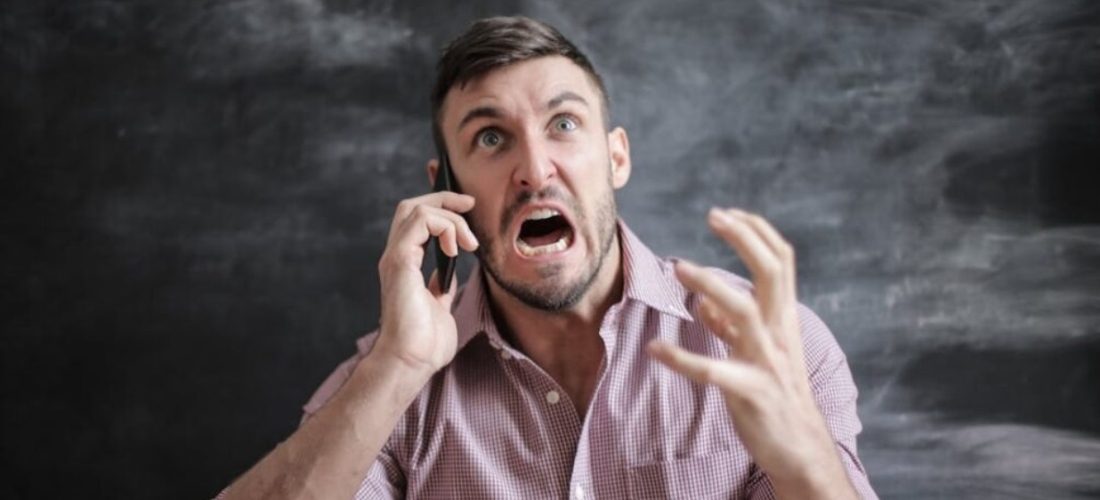 A man in a checkered shirt speaks on the phone, gesturing expressively against a dark background.